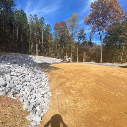 Excavated site with gravel and machinery, surrounded by trees under a blue sky.