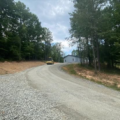 Gravel driveway leading to a gray building surrounded by trees under a cloudy sky.