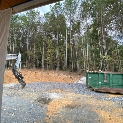 Construction site with excavator and green dumpster, surrounded by trees and gravel.
