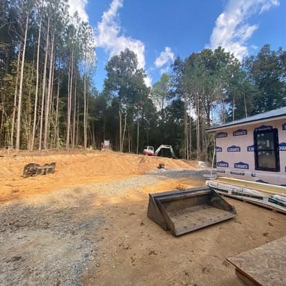 Construction site showing cleared land, trees, and building materials under a blue sky.