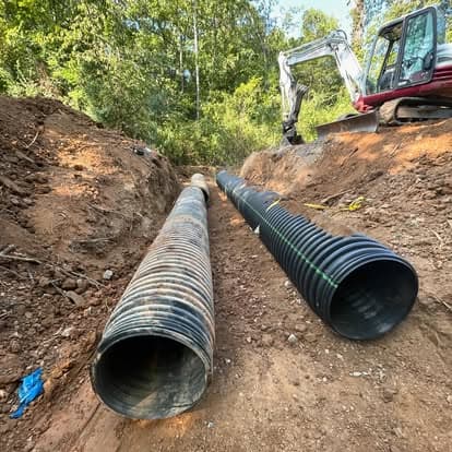 Excavation site showing two large drainage pipes and equipment among trees.