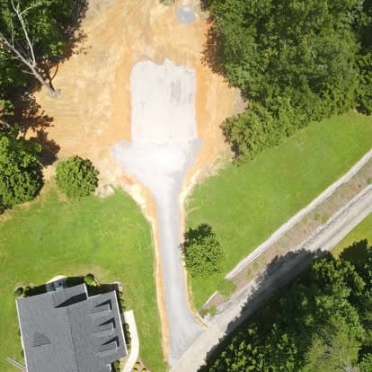 Aerial view of a cleared lot with a gravel driveway, surrounded by greenery and trees.