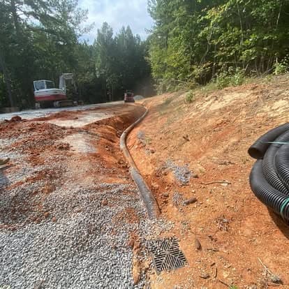 Construction site with drainage pipe installation and gravel surface, surrounded by trees.