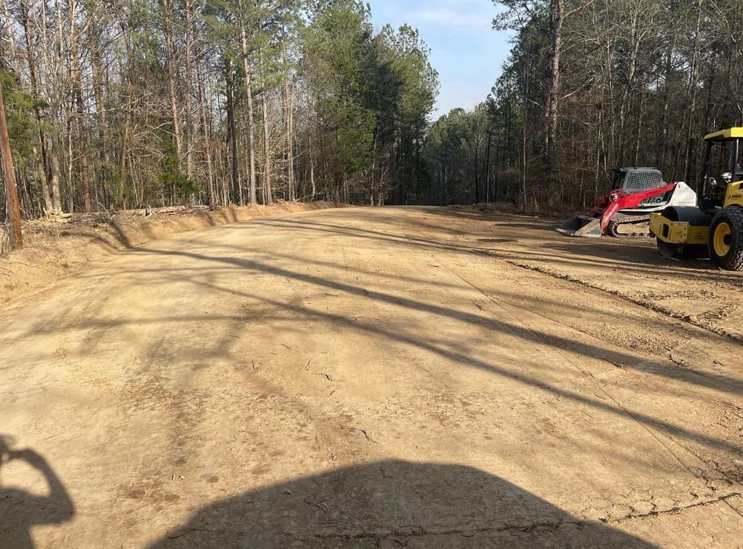 Cleared dirt road in forest area with construction equipment and trees along the sides.