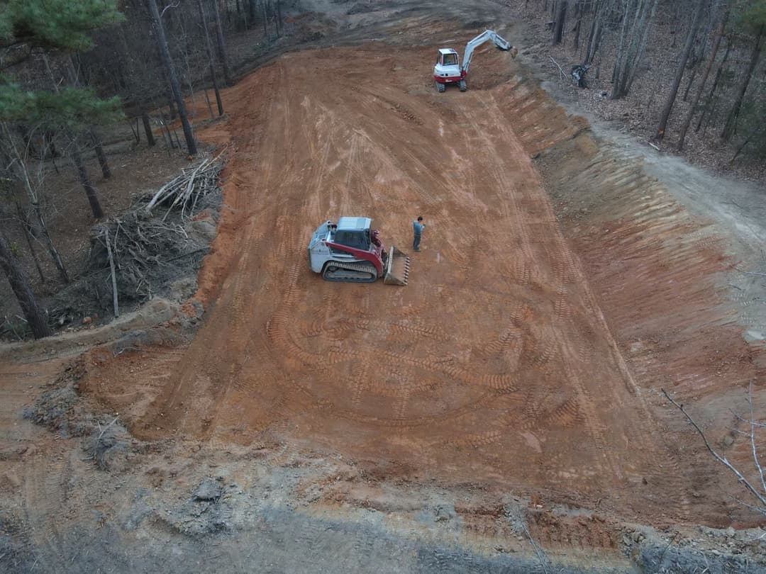 Construction site with heavy machinery and a worker on freshly cleared dirt in a wooded area.